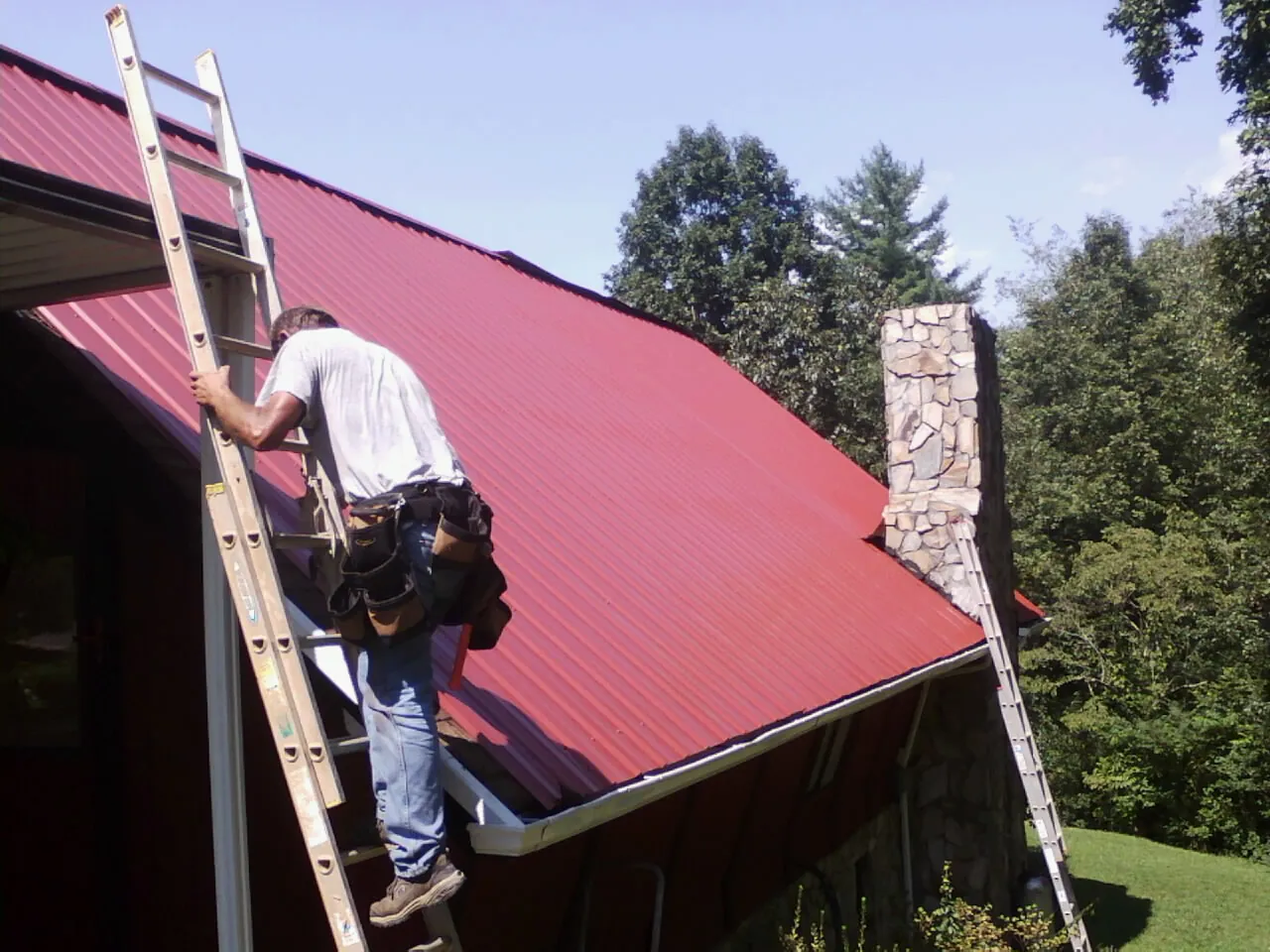 Roofer on a ladder beside a red metal roof and stone chimney — Soffit & Fascia Repair by Roofing Brothers in Mehama